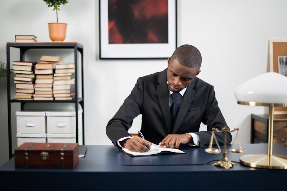 Businessman in formal suit writing notes at office desk with books and scales of justice.